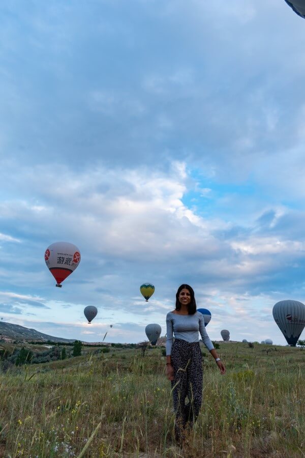 woman in grey top and long flowy pants posing in front of hot air balloons on a green field in Cappadocia