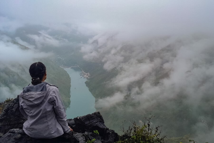 a woman in a grey raincoat sitting at a viewpoint overlooking nho que river in ha giang