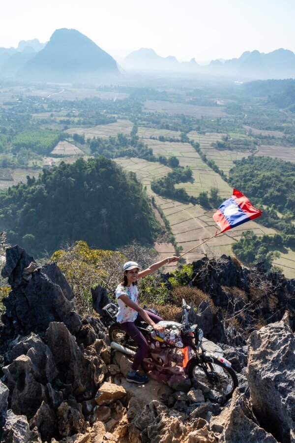 a woman posing on an old motorbike while waving a laos flag on a mountain in vang vieng